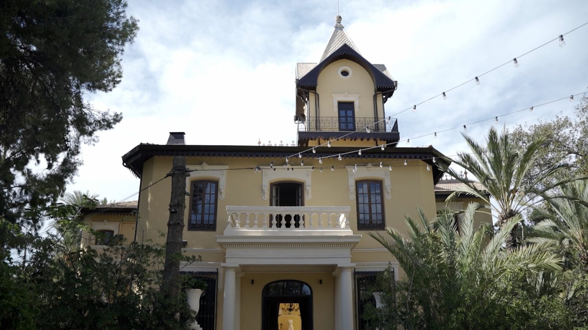 Casa antigua amarilla con torre y balcones rodeada de árboles y luces colgantes en exterior