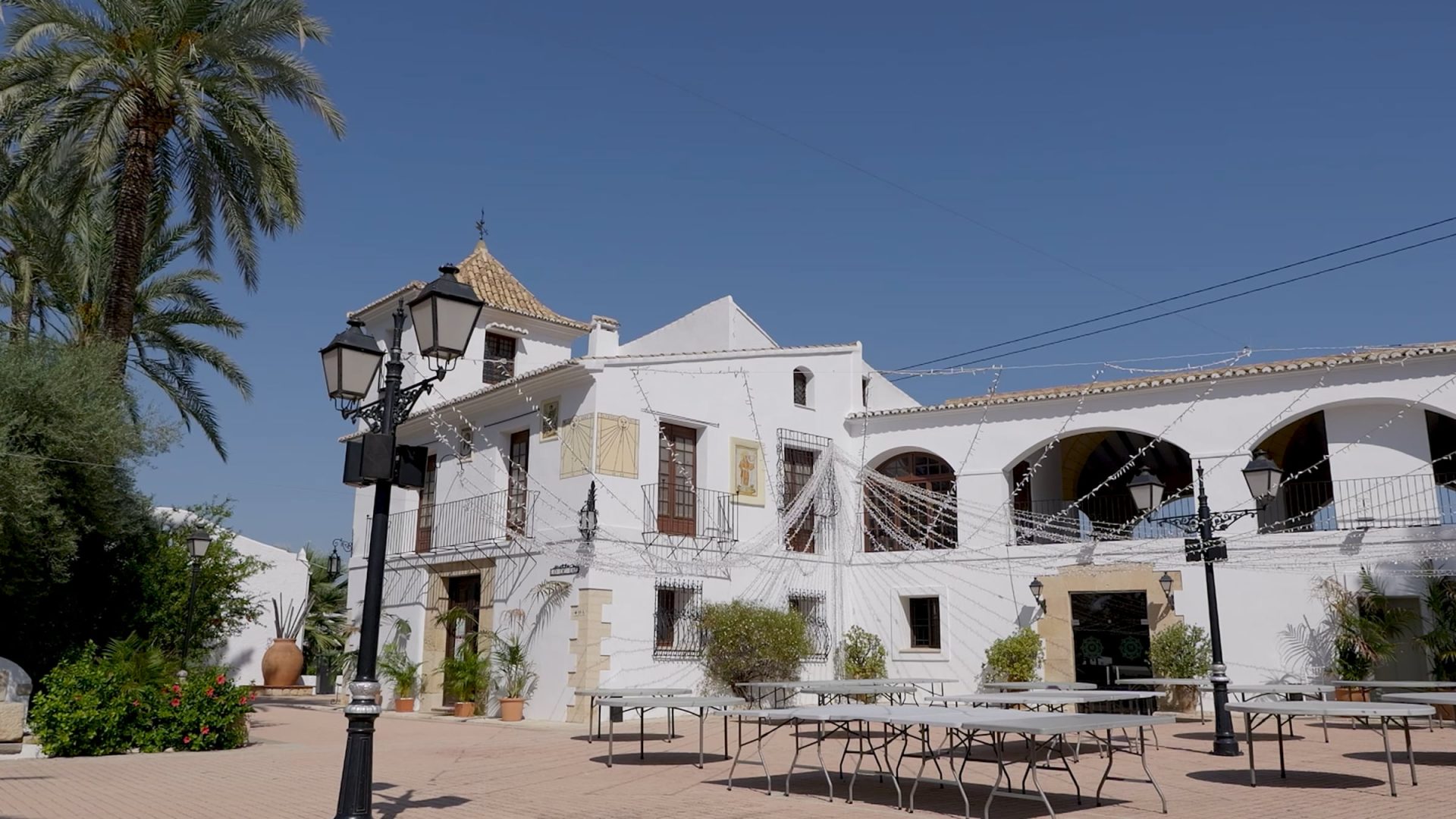 plaza exterior con farolas, mesas plegables y edificio blanco colonial bajo cielo azul